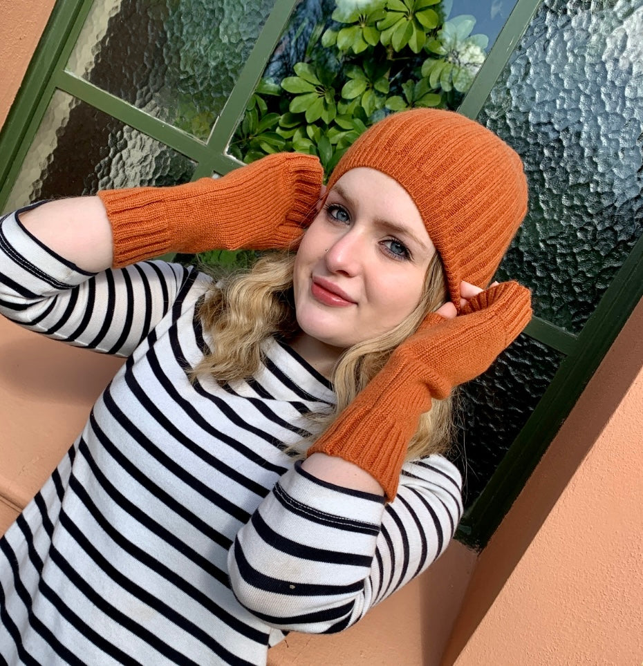A woman wearing a BROOME baby rib cashmere knit beanie in treacle and matching treacle GOTTA HAND IT TO YOU cashmere fingerless gloves, paired with a black and white striped long-sleeve top, posing in front of a window.