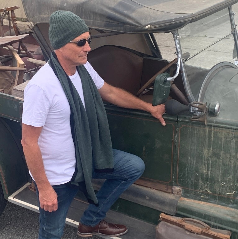 A man wearing a BROOME pine green baby rib knit cashmere beanie, sunglasses, a white t-shirt, and a I'M WRAPPED pine green knit cashmere scarf stands next to an antique green vehicle.