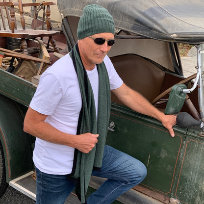 A man wearing a BROOME pine green baby rib knit cashmere beanie, sunglasses, a white t-shirt, and a I'M WRAPPED pine green knit cashmere scarf stands next to an antique green vehicle.