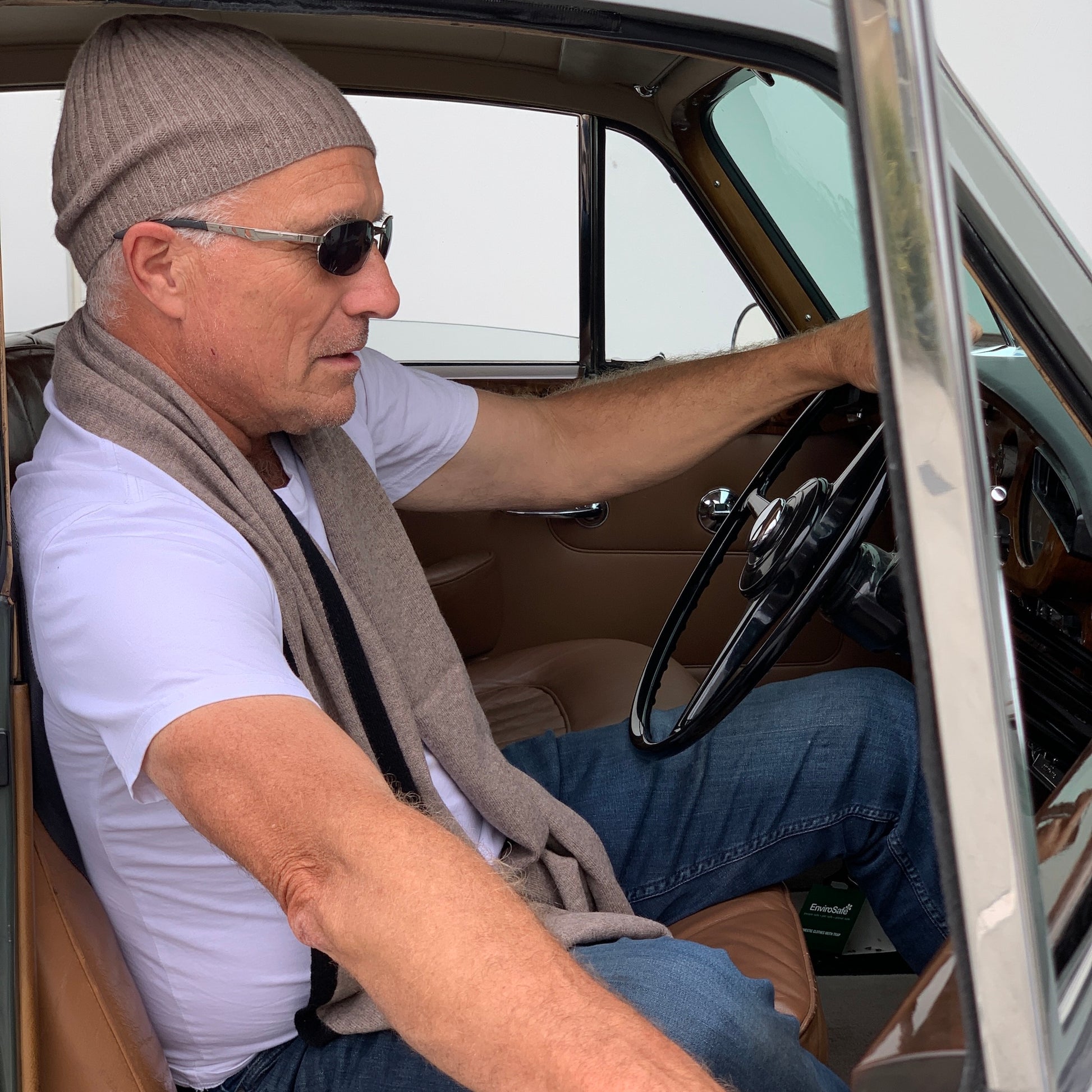 A man wearing a BROOME donkey brown baby rib knit cashmere beanie, sunglasses, a white t-shirt, and a I'M WRAPPED donkey brown knit cashmere scarf sitting in an antique green car.