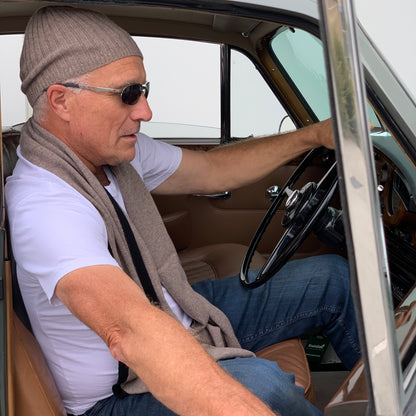 A man wearing a BROOME donkey brown baby rib knit cashmere beanie, sunglasses, a white t-shirt, and a I'M WRAPPED donkey brown knit cashmere scarf sitting in an antique green car.