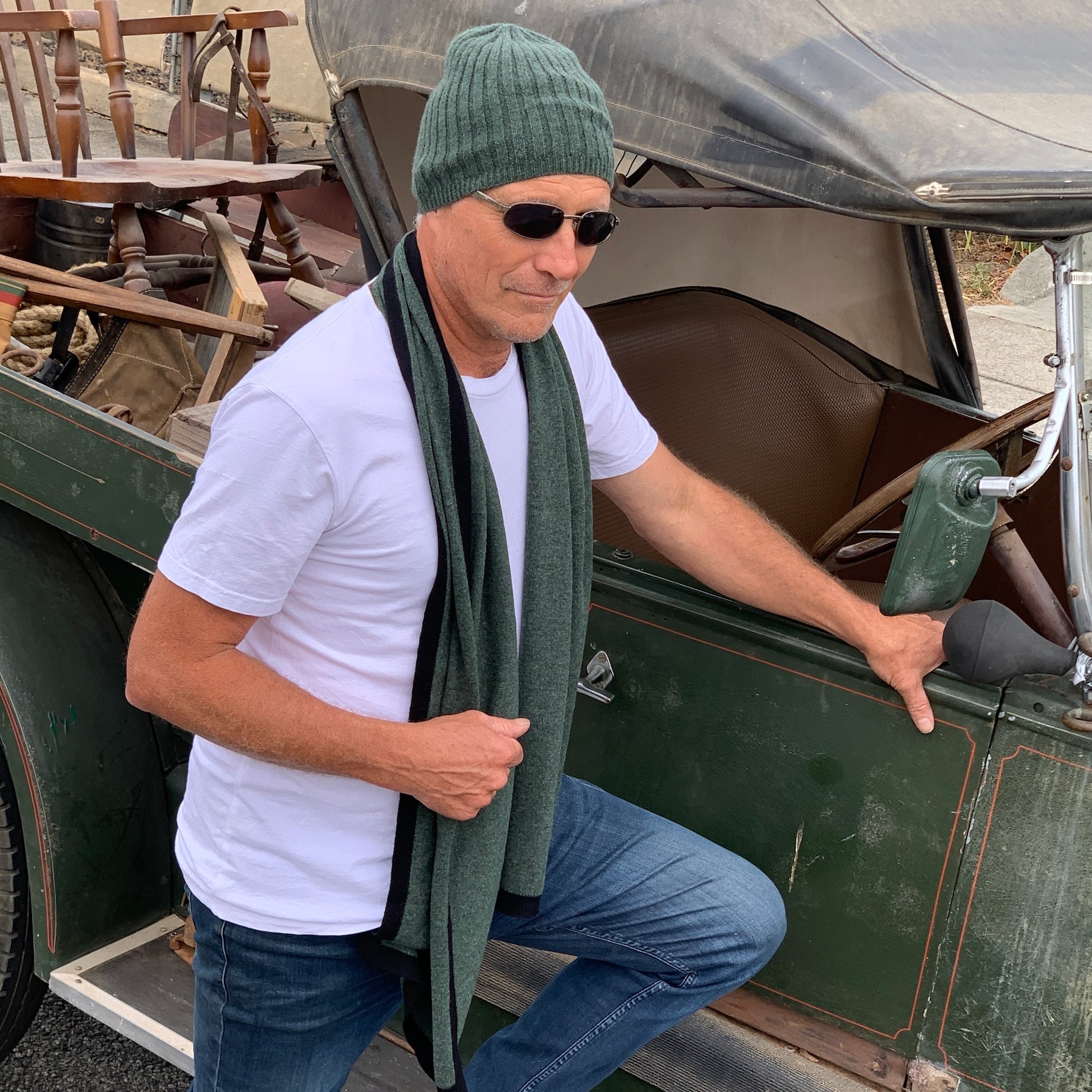 A man wearing a BROOME pine green baby rib knit cashmere beanie, sunglasses, a white t-shirt, and a I'M WRAPPED pine green knit cashmere scarf stands next to an antique green vehicle.